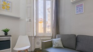 A room with a grey sofa, a white chair, a radiator, a window with a view of buildings, and a plant on a shelf. - Conte Verde - Bilo 7 Grey by Wonderful Italy (Turin)