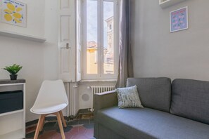 A room with a grey sofa, a white chair, a radiator, a window with a view of buildings, and a plant on a shelf. - Conte Verde - Bilo 7 Grey by Wonderful Italy (Turin)