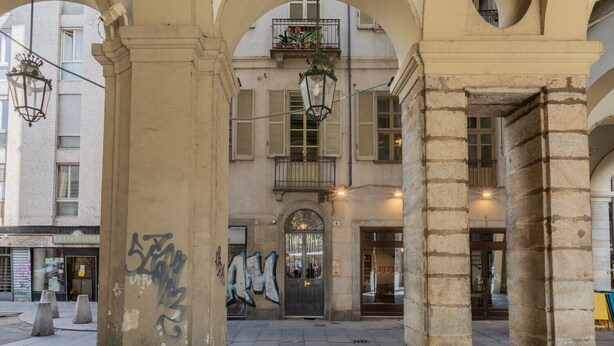 An urban street scene with a building facade, a door with graffiti, and hanging street lamps. - Conte Verde - Bilo 7 Grey by Wonderful Italy (Turin)