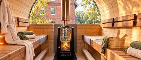 A sauna room with wooden benches, a central wood-burning stove, and a view through a semi-circular window.