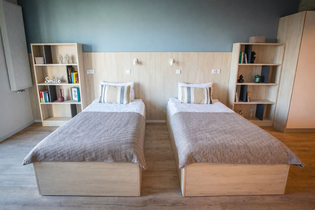 Une chambre avec deux lits, des têtes de lit en bois, une bibliothèque et un luminaire fixé au mur.