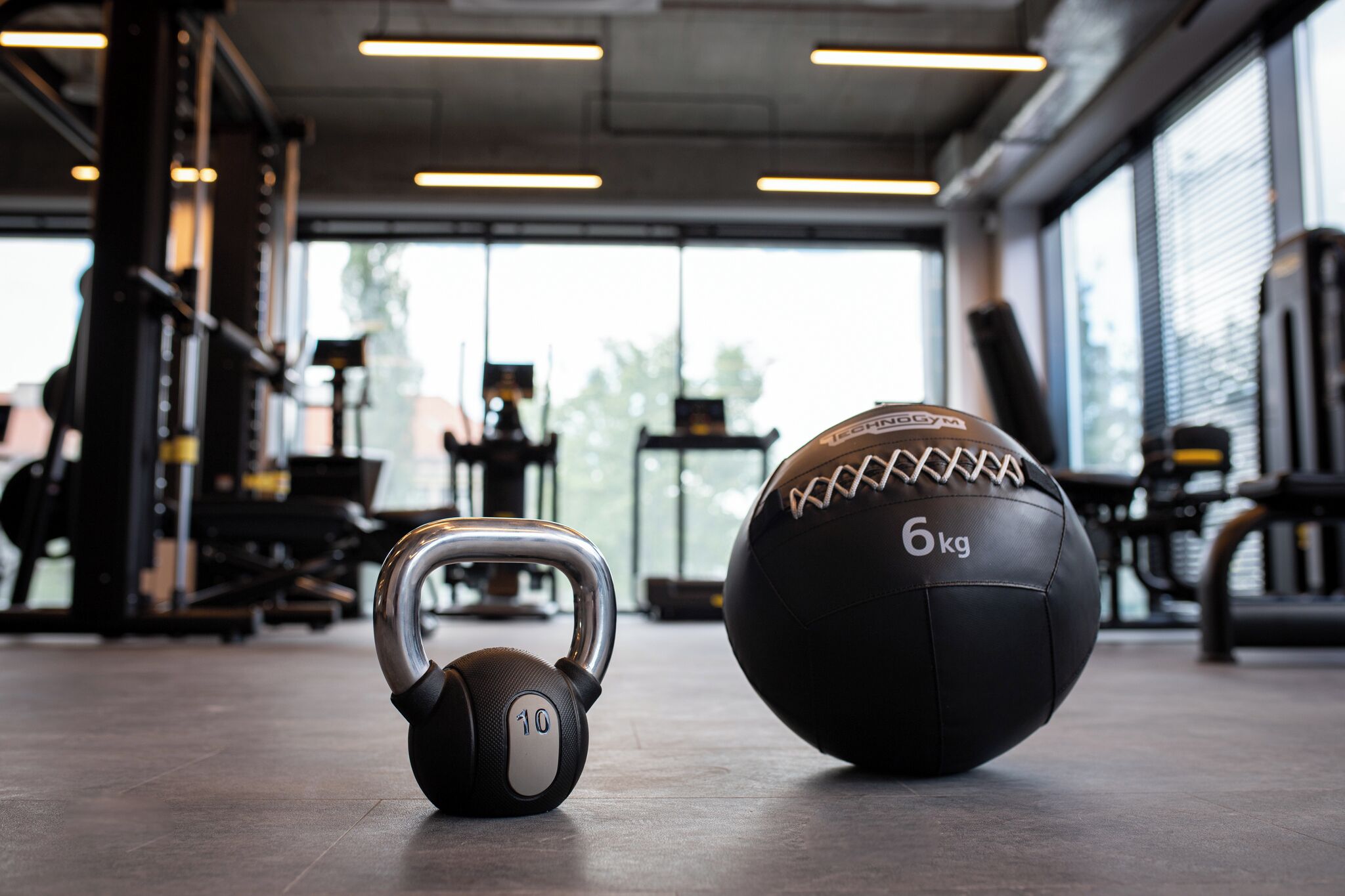 A kettlebell and a medicine ball in a gym with exercise equipment and large windows.