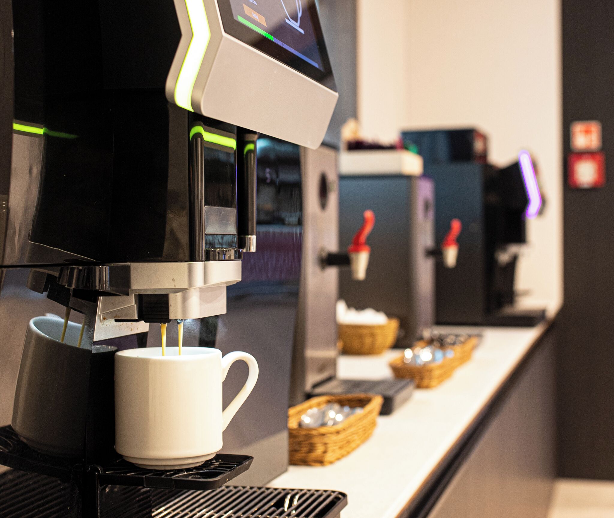 Una máquina de café sirviendo café en una taza, con otras máquinas y cestas al fondo.