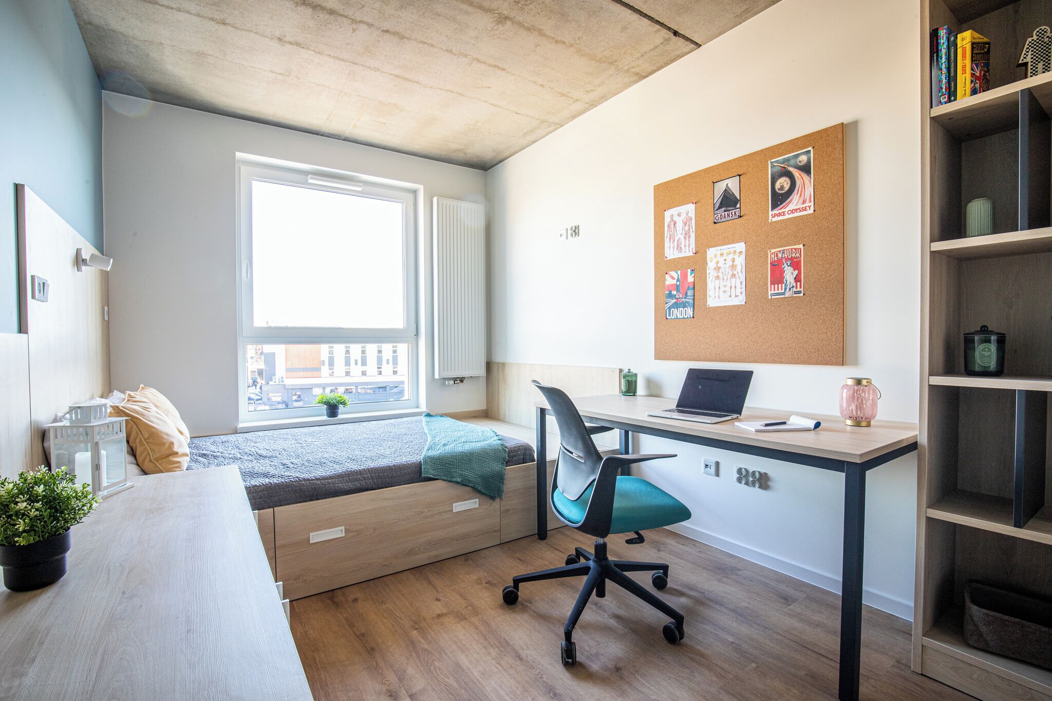 Una habitación con cama, escritorio, un tablón de anuncios con fotografías, ventana y vistas a edificios.