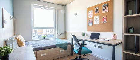 Una habitación con cama, escritorio, un tablón de anuncios con fotografías, ventana y vistas a edificios.