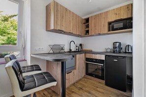 A compact kitchen with wooden cabinets, a built-in oven, a dishwasher, and a window with a view of greenery. - Apartment "Aurore" with Wi-Fi in Cancale, 1st Floor (Cancale)