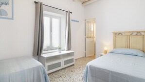 Un dormitorio con dos camas, una ventana con cortinas, una mesita de noche con lámpara y una puerta que da a otra habitación.