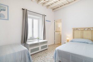 Quarto com duas camas, janela com cortinas, mesa de cabeceira com abajur e porta para outro cômodo.