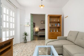 Quarto com sofá, armário de madeira, mesa de centro de vidro, planta em vaso e porta que dá para outro cômodo.