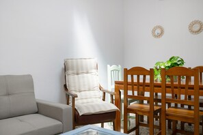 Uma sala de estar com sofá, poltrona acolchoada, mesa de jantar de madeira com cadeiras e um vaso de planta.