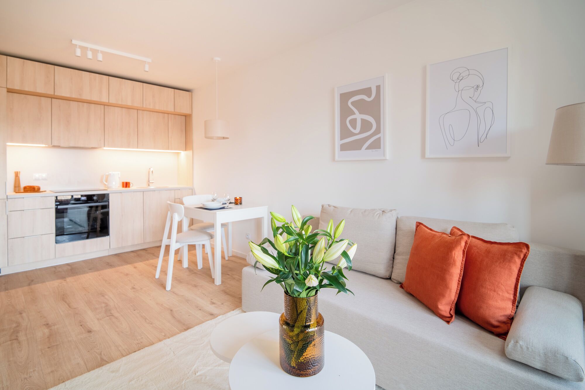 A modern kitchen with wooden cabinets, a dining area with a white table and chairs, a living room with a sofa and cushions, and a vase with flowers on a side table.