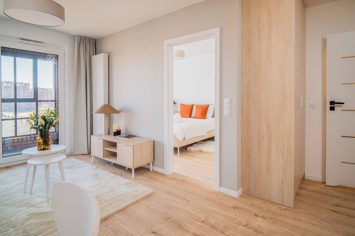 A modern hotel room with a large window, a desk, a bed with orange pillows, and a wooden door.
