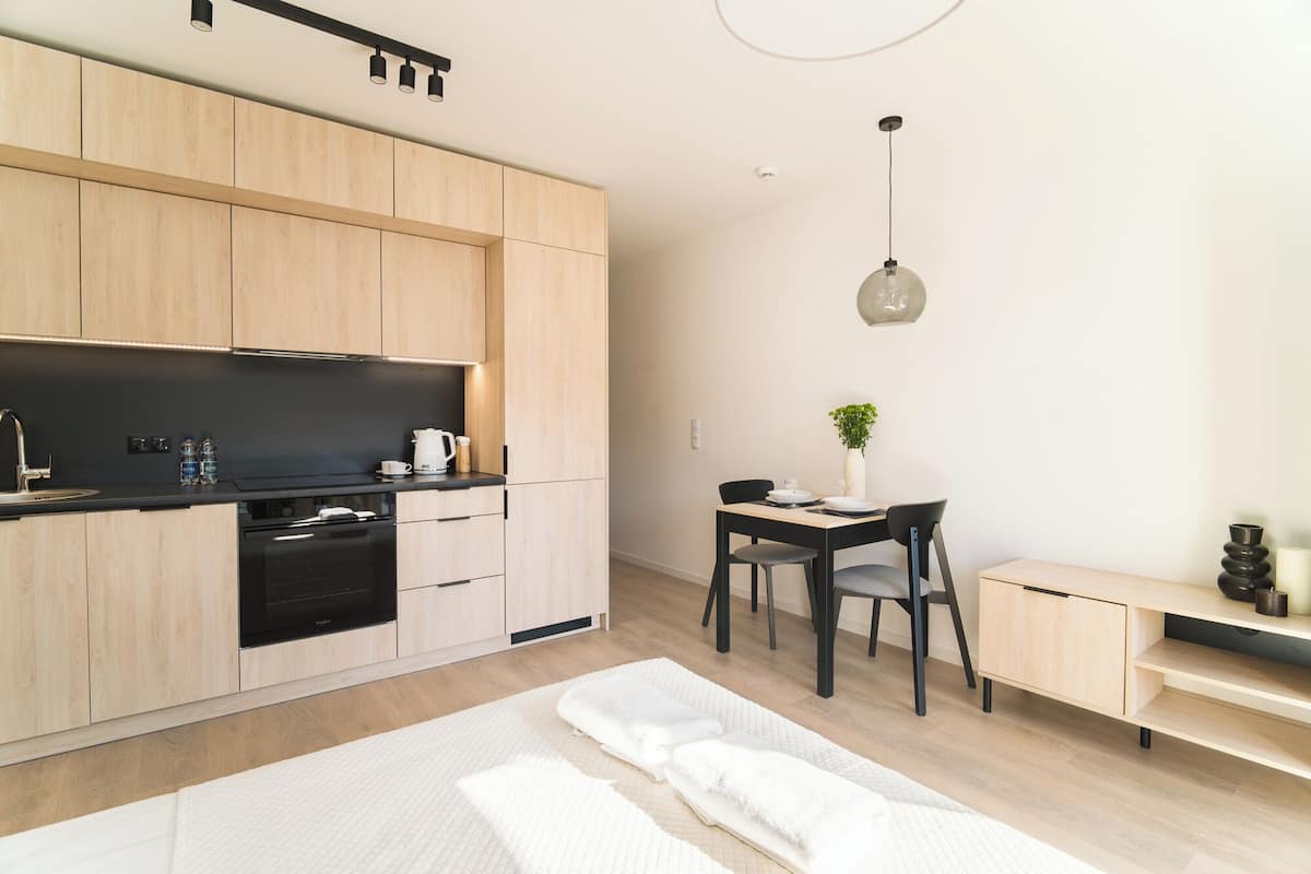 A modern kitchen with wooden cabinets, a black countertop, and a built-in oven. There is a dining area with a table, chairs, and a vase with a plant.