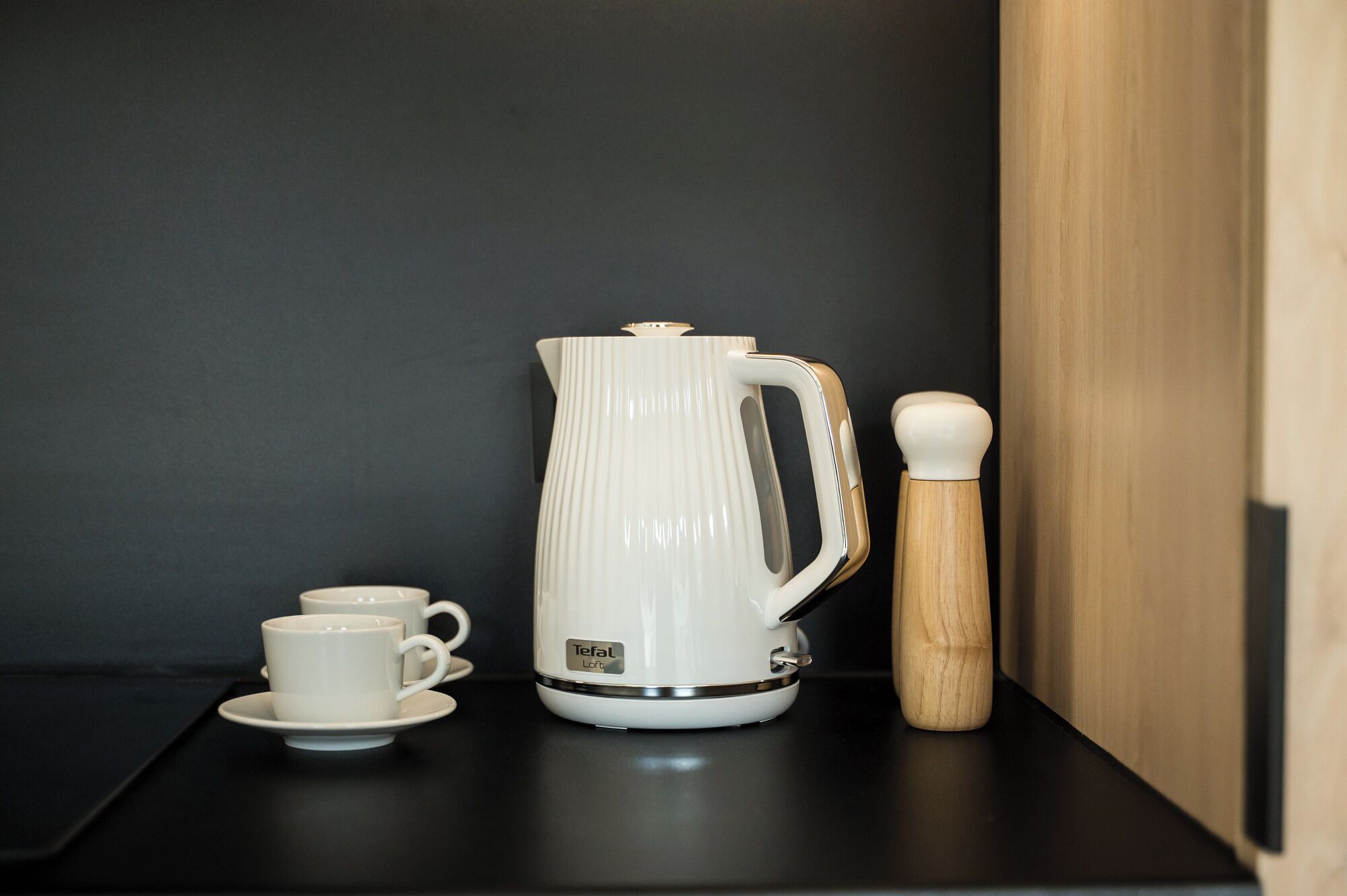 Una tetera eléctrica blanca con asa y pico, una taza blanca sobre un platito, y un molinillo de pimienta de madera sobre una encimera negra.