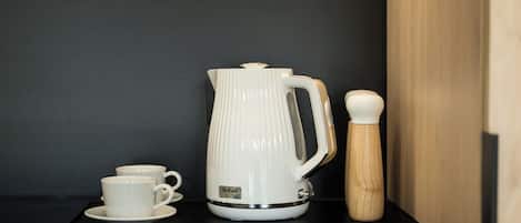 Una tetera eléctrica blanca con asa y pico, una taza blanca sobre un platito, y un molinillo de pimienta de madera sobre una encimera negra.