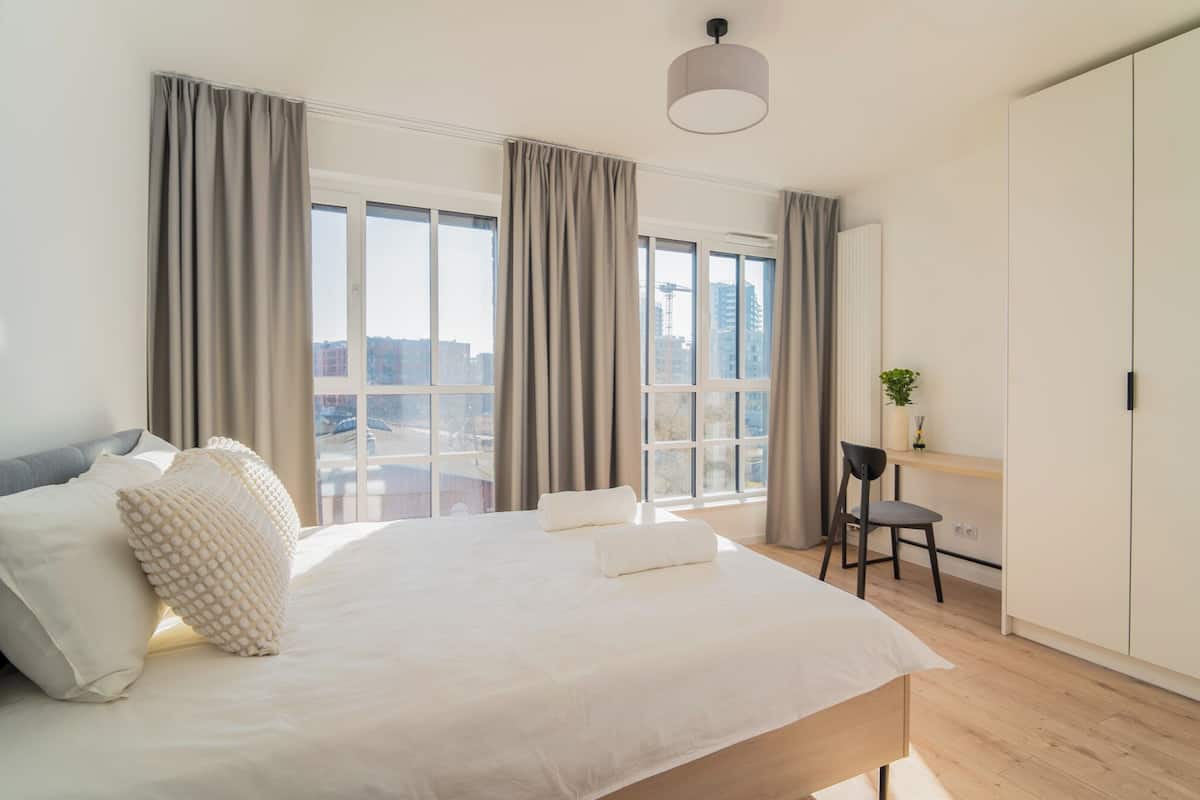 A modern bedroom with a large bed, a desk, and a chair, all with a view of the city through large windows.