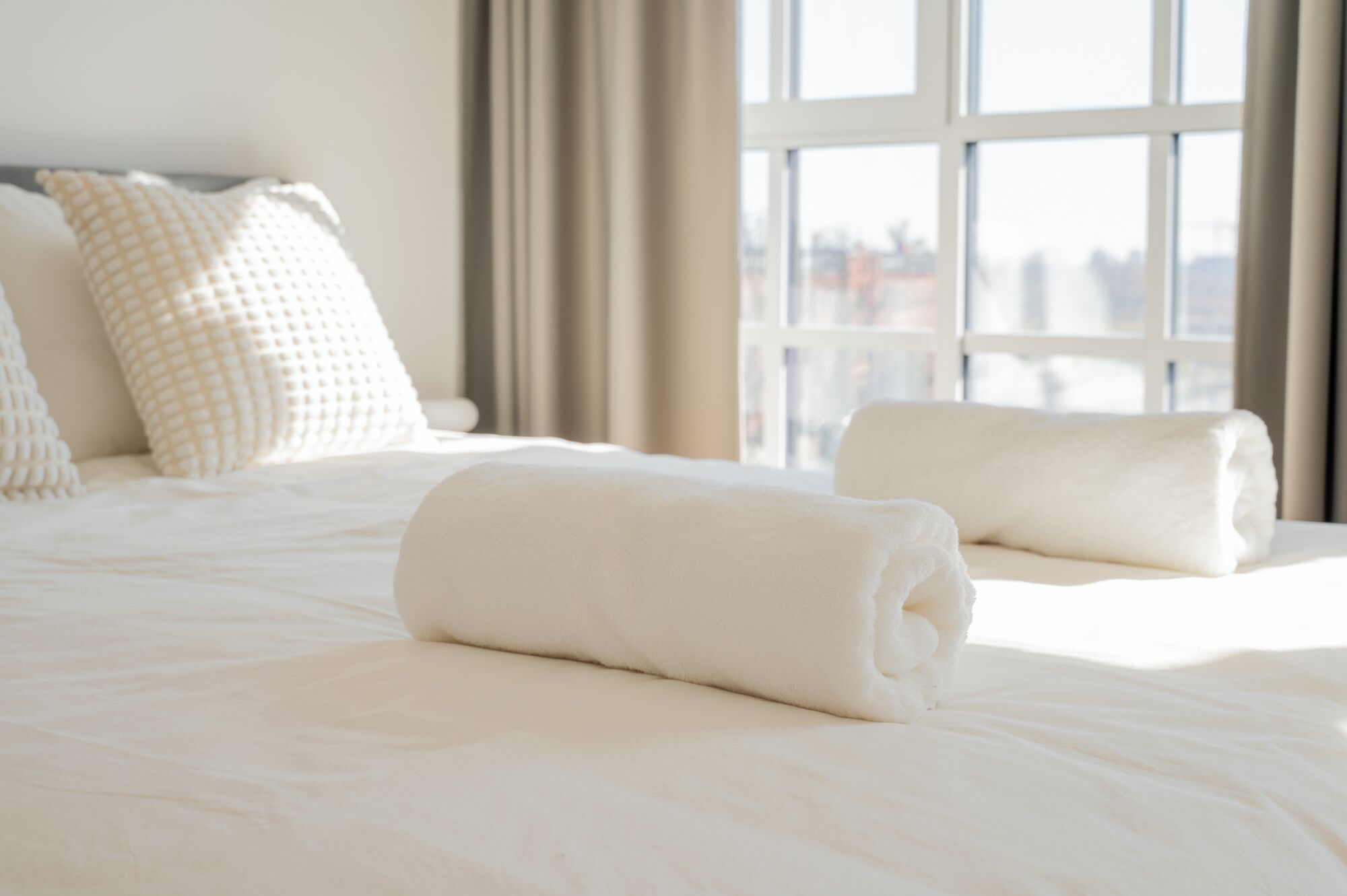 A neatly made bed with white linens and pillows, a window with a view of buildings, and a curtain.