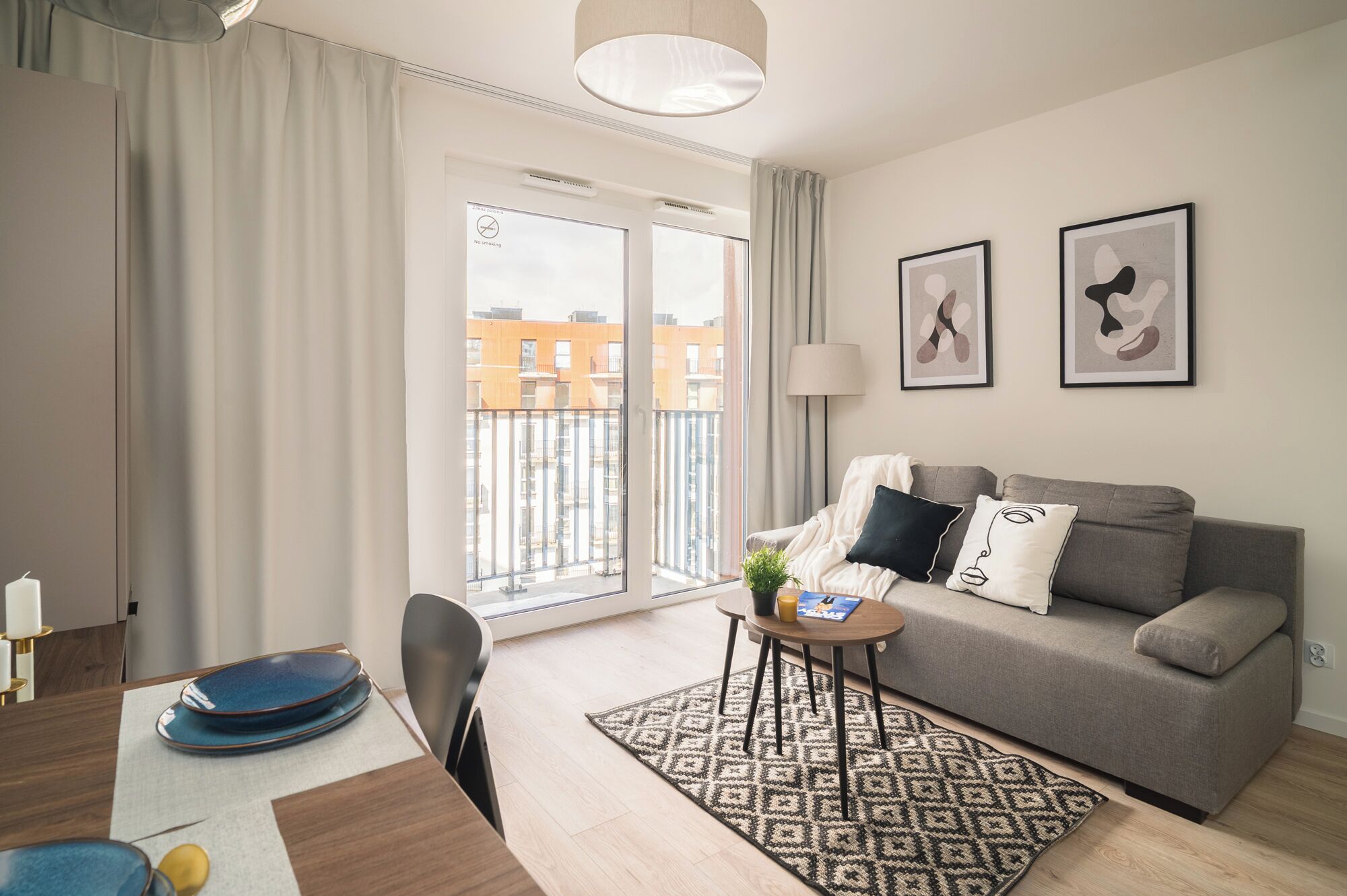 A modern living room with a sofa, a small table, a lamp, and a view of buildings through a large window.