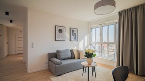 A modern living room with a grey sofa, a round table with a vase of flowers, and a large window with curtains. - Resi Apartments (Gdańsk)
