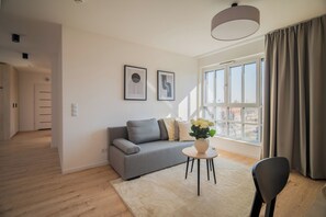 Un salon moderne avec un canapé gris, une table ronde ornée d’un vase de fleurs et une grande fenêtre avec des rideaux.