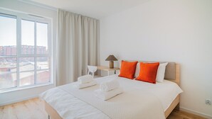 A hotel room with a large bed, a desk, a lamp, and a view of buildings outside. - Resi Apartments (Gdańsk)