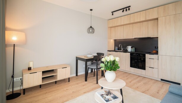 A modern kitchen with wooden cabinets, a dining table with chairs, a small round table with a vase of flowers, and a floor lamp. - Resi Apartments (Gdańsk)