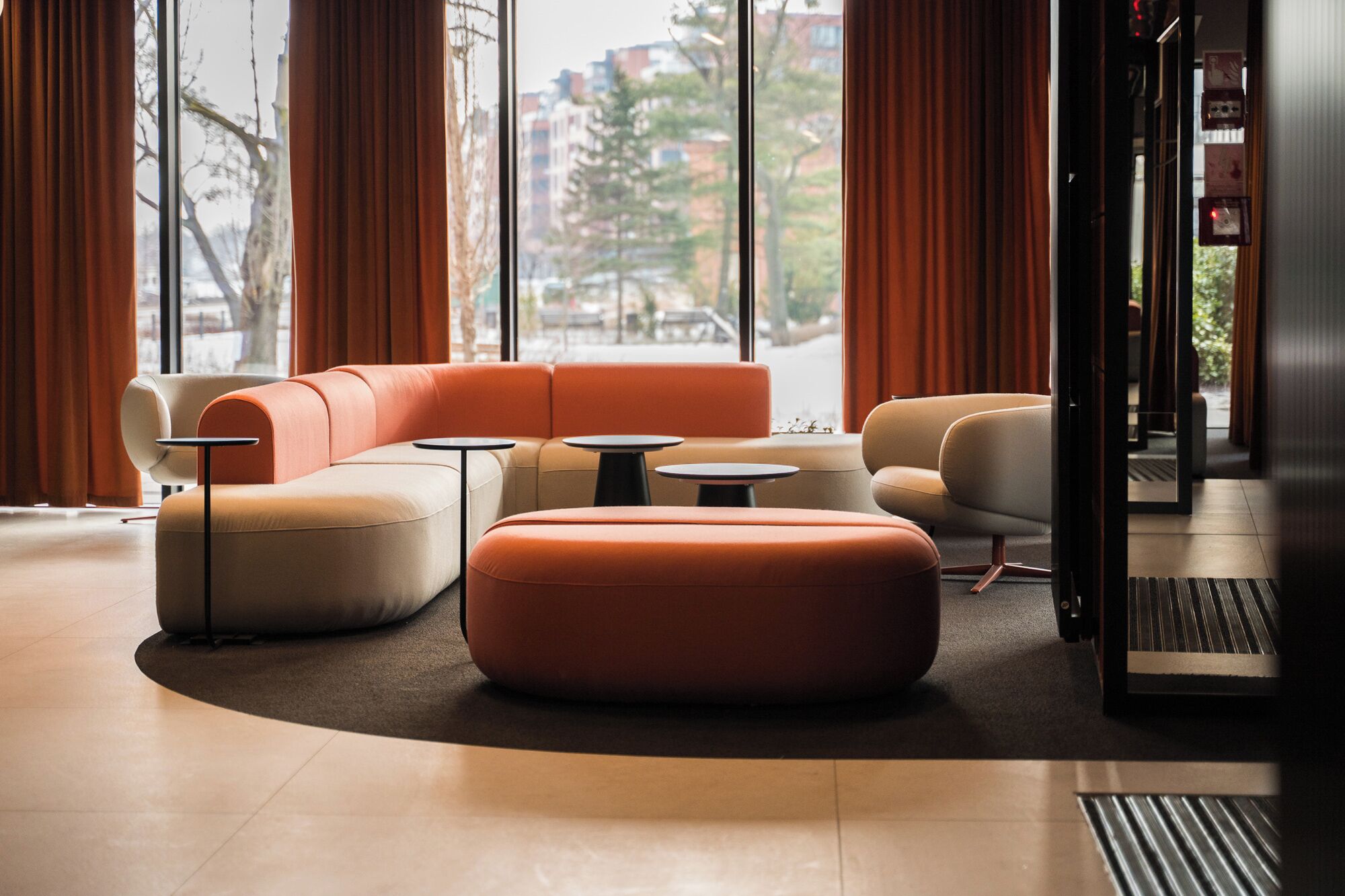 Un espace de détente moderne comprenant un canapé, un pouf et de petites tables, de grandes fenêtres avec des rideaux et une vue dégagée sur le paysage urbain.