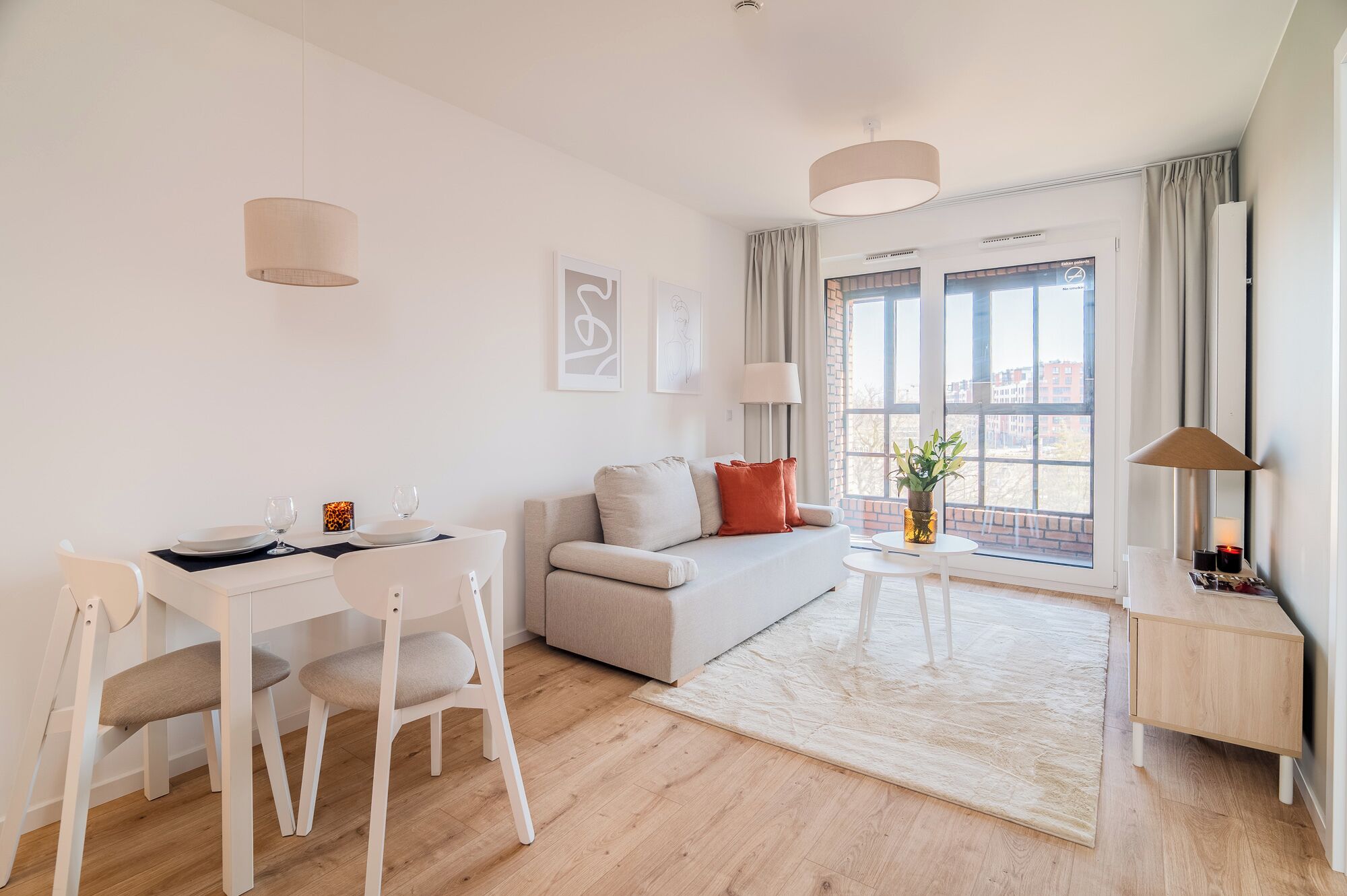 A modern living room with a sofa, a dining table with chairs, a small side table with a lamp, and a large window with a view of buildings.