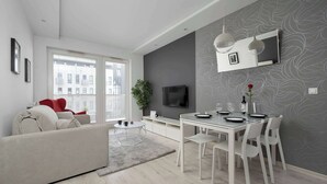 Een moderne woonkamer met een bank, een eettafel en stoelen, een flatscreen televisie aan de muur en een groot raam met uitzicht op gebouwen.