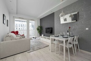 Un salon moderne comprenant un canapé, une table à manger et des chaises, un téléviseur à écran plat fixé au mur et une grande fenêtre donnant sur des immeubles.