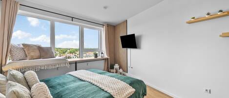 A bedroom with a large bed, a window with a view, a mounted TV, and a shelf with plants.