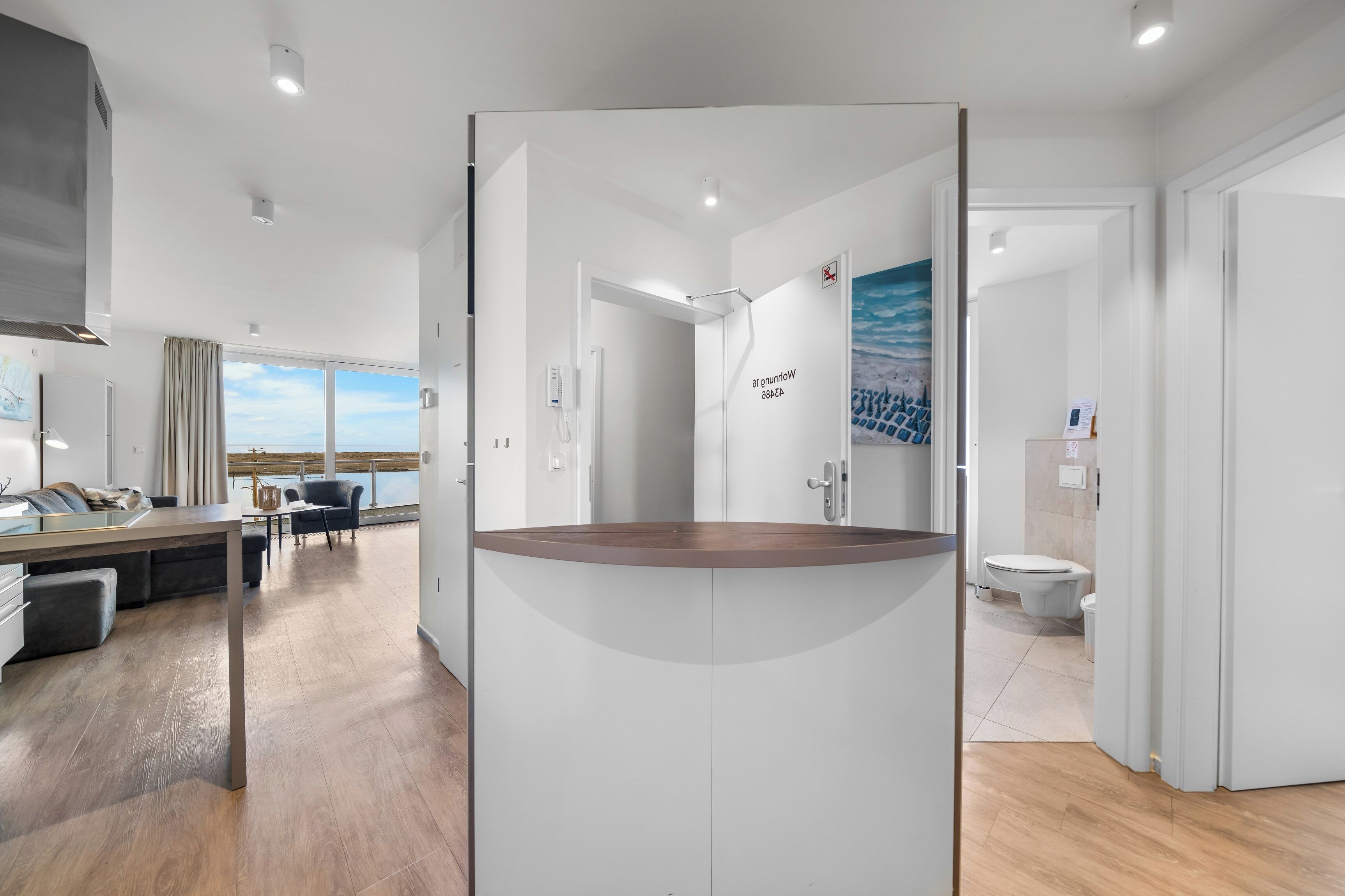 A modern hotel room with a wooden floor, a round table, a sofa, a view of the sea, and a bathroom with a toilet and sink.