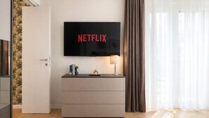 Ein modernes Hotelzimmer mit einem wandmontierten Flachbildfernseher, einem Nachttisch mit Lampe und einem großen Fenster mit Vorhängen.