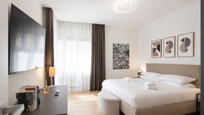 Ein modernes Hotelzimmer mit einem großen Bett, einem Flachbildfernseher, einem Schreibtisch mit Büchern und einer Lampe sowie Kunstwerken an den Wänden.