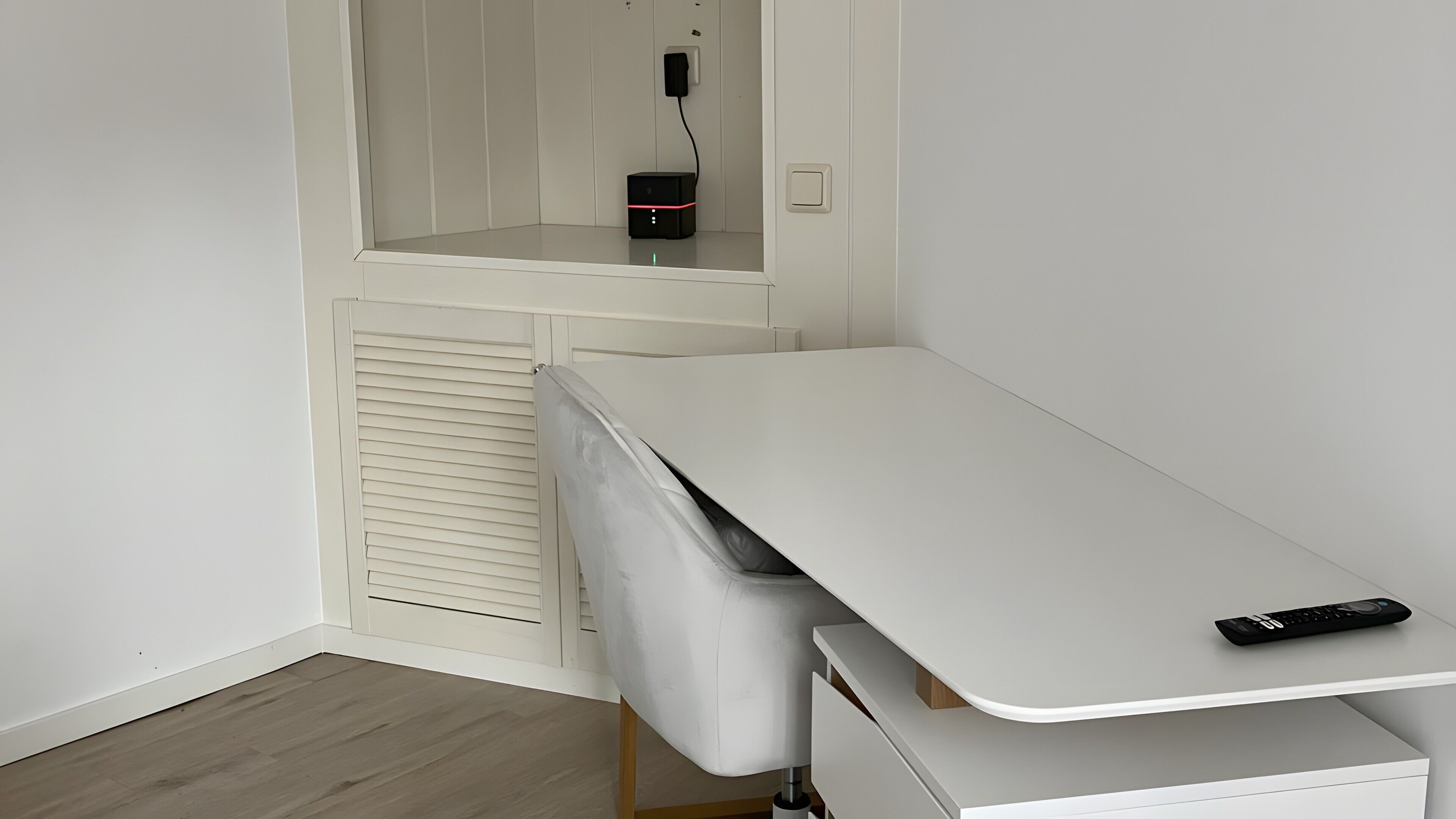 Ein Zimmer mit weißem Schreibtisch, einem Stuhl, einem kleinen schwarzen Gerät auf dem Schreibtisch und einem weißen Schrank mit Spiegel.
