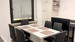 Une pièce comprenant une table d’échecs, des chaises, une fenêtre avec des stores, une armoire blanche et une petite étagère.