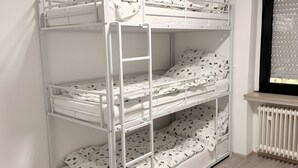 Un lit superposé avec une structure blanche et une literie à motifs, une fenêtre avec des stores et un radiateur en dessous.