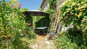 Un jardin avec une table et une chaise sous une tonnelle, entouré d’une végétation luxuriante et de plantes en fleurs.