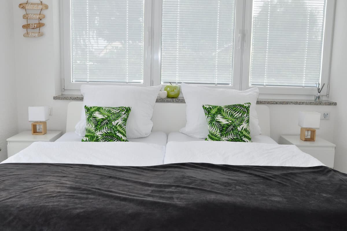 Una cama bien hecha con ropa blanca y almohadas estampadas con hojas, una mesita de noche con lámpara, y una ventana con persianas.