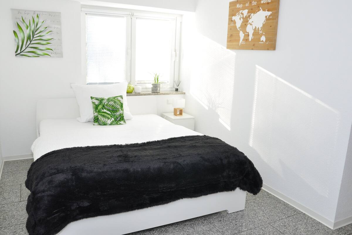 Un dormitorio con cama, ventana, mesita de noche con lámpara, un mapa del mundo en la pared y una planta en el alféizar.