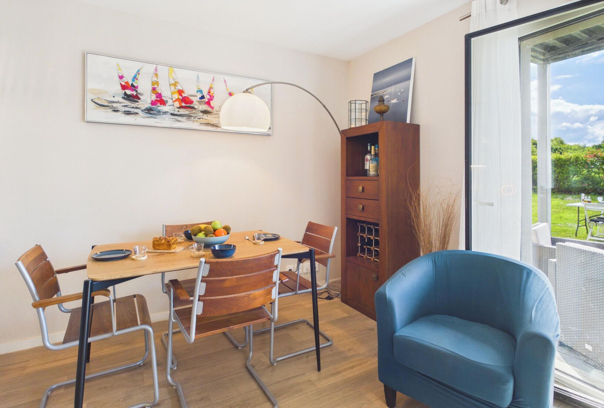 A dining area with a wooden table, chairs, a blue armchair, a painting, and a view through a glass door.