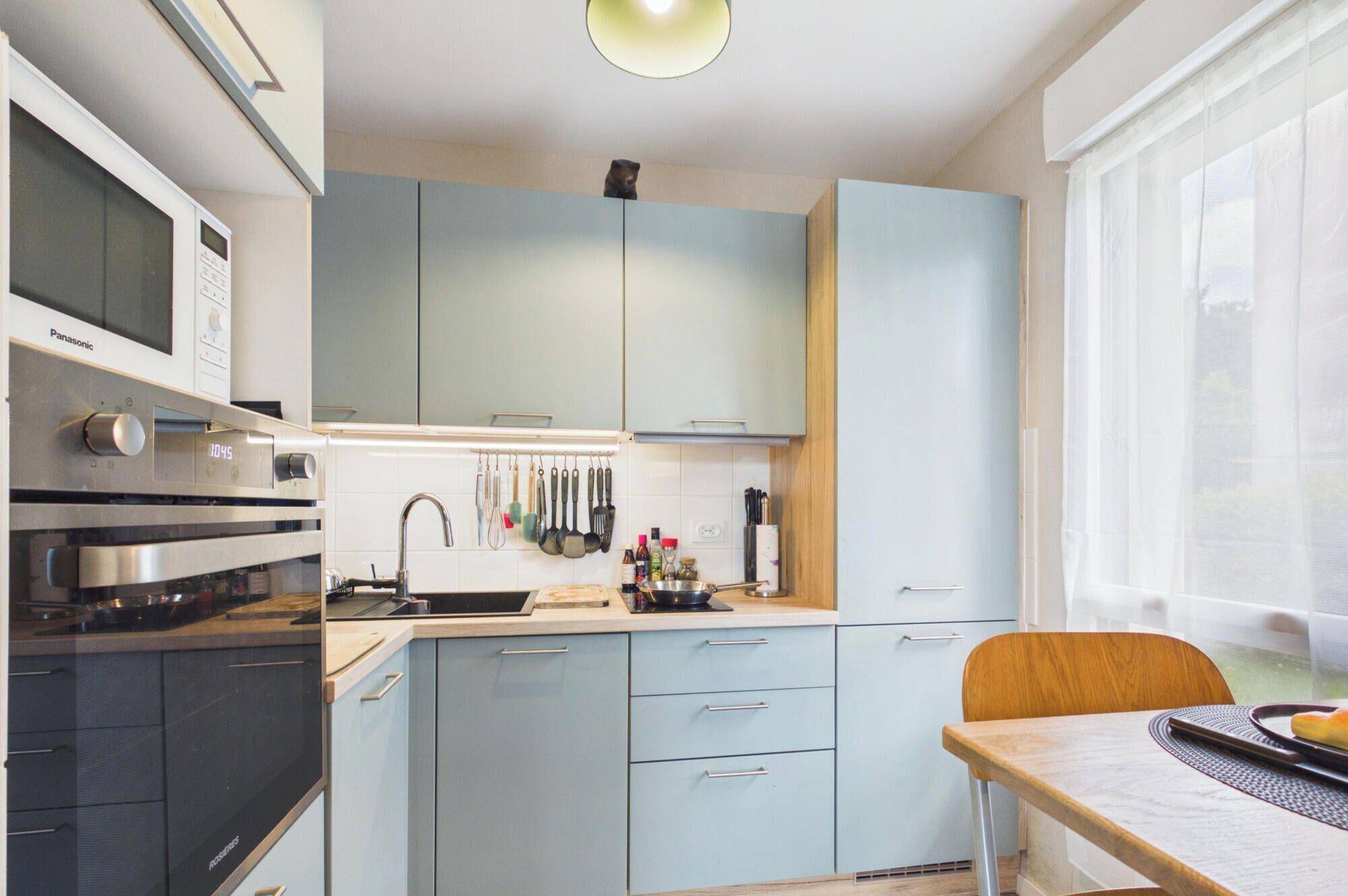 Eine moderne Küche mit Mikrowelle, Backofen und Spüle. Ein Fenster mit Vorhängen und ein Esstisch mit einem Stuhl sind ebenfalls vorhanden.