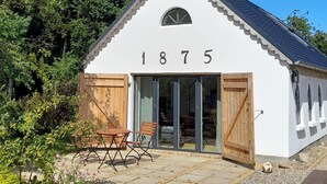Een wit gebouw met het nummer 1875, houten deuren, een patio met een tafel en stoelen, en een tuin.