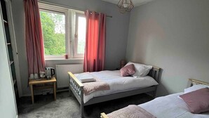 A bedroom with two beds, a radiator, a table with cups and a kettle, and a window with curtains. - Room-amina's Cottage Women Only Accommod (Gloucester)