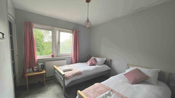 A bedroom with two single beds, a window with curtains, a radiator, and a small table with a kettle. - Room-amina's Cottage Women Only Accommod (Gloucester)