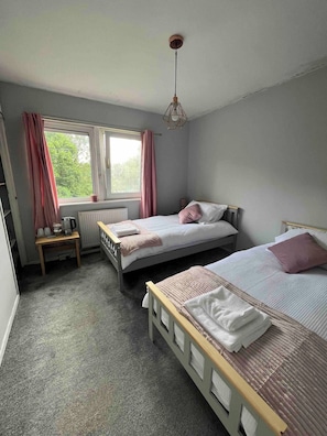 A bedroom with two beds, a window with curtains, a radiator, and a table with a kettle. - Room-amina's Cottage Women Only Accommod (Gloucester)