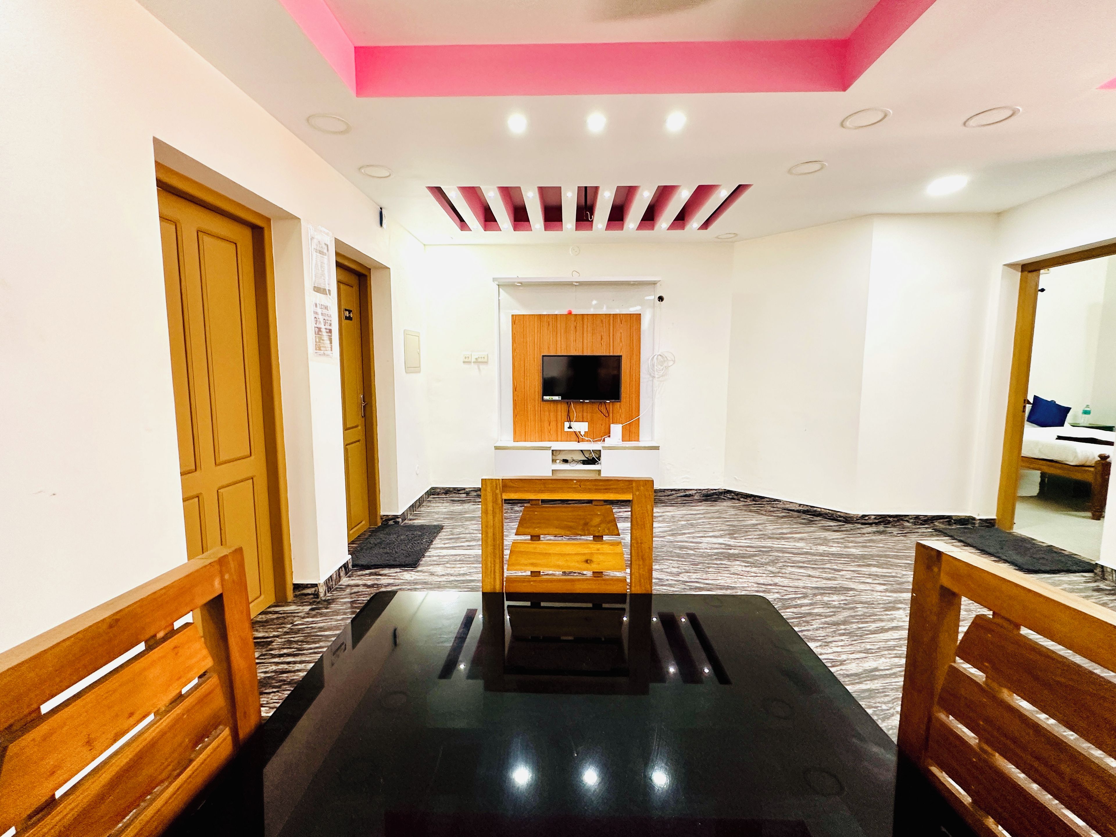 Habitación de hotel con una mesa de comedor de madera, un televisor de pantalla plana en la pared y un techo con iluminación empotrada.