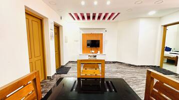 Habitación de hotel con una mesa de comedor de madera, un televisor de pantalla plana en la pared y un techo con iluminación empotrada.