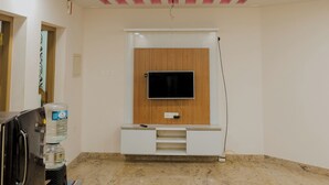 A room with a flat-screen TV mounted on a wooden panel, a ceiling fan, recessed lighting, and a water dispenser. - Varkala Breeze Villas (Varkala)
