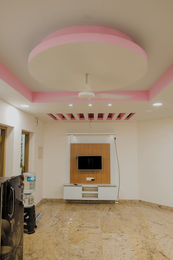 Habitación con televisión de pantalla plana montada en un panel de madera, ventilador de techo, luces empotradas y dispensador de agua.